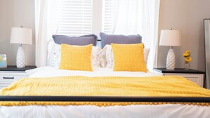 A neatly made bed with a yellow blanket, two pillows with a textured pattern, and a white headboard. There are two bedside tables with lamps and a small vase with flowers. - The Bluebonnet - Pet-Friendly Historic 3BR Home in Downtown McKinney (McKinney)