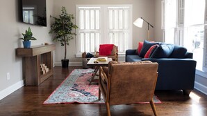A living room with a dark blue sofa, a wooden coffee table, a red chair, a large plant, and a patterned rug. - The Bluebonnet - Pet-Friendly Historic 3BR Home in Downtown McKinney (McKinney)