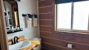 Un baño pequeño con un tocador de madera, un lavamanos blanco, un inodoro y una ventana con barandilla de cuerda.