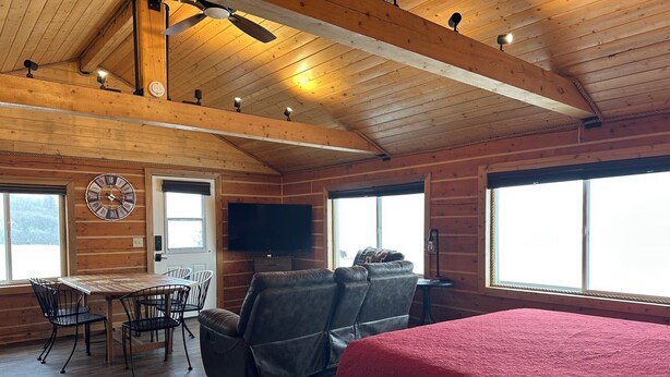 Un acogedor interior de cabaña con techo de madera, una cama grande con manta roja, un sofá, un televisor y un comedor con sillas y una mesa.