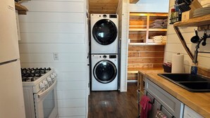 Una cocina compacta con estufa a gas, dos lavadoras apiladas, un estante de madera con toallas y un lavabo con una botella de detergente.
