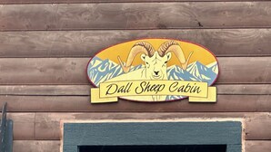 A wooden cabin with a sign that reads "Dall Sheep Cabin" featuring a mountain and ram illustration. - Dall Cabin - Cozy retreat with bay views, a shared hot tub, and fire pit! (Homer)