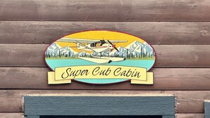 Eine Holzhütte mit einem Schild, auf dem 'Super Cut Cabin' steht, mit einem Flugzeug und Bergen im Hintergrund.
