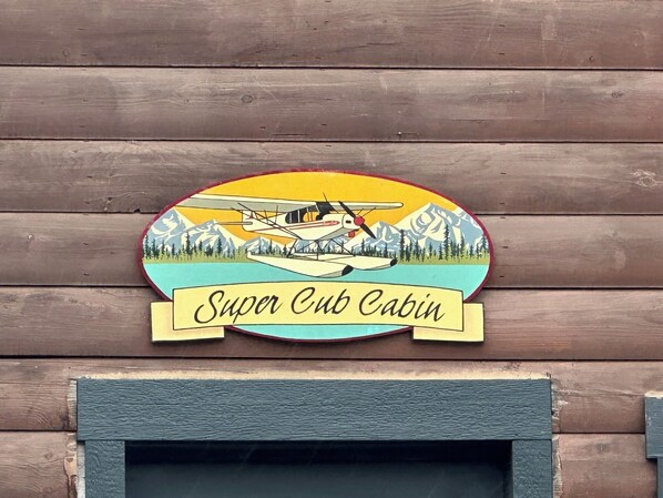 Una cabaña de madera con un letrero que dice "Super Cut Cabin", con un avión y montañas.