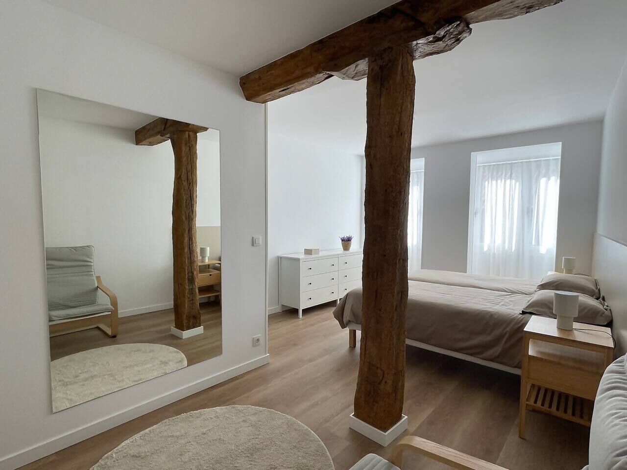 Quarto com um espelho grande, vigas de madeira, uma cama, cômoda e janela com cortinas.