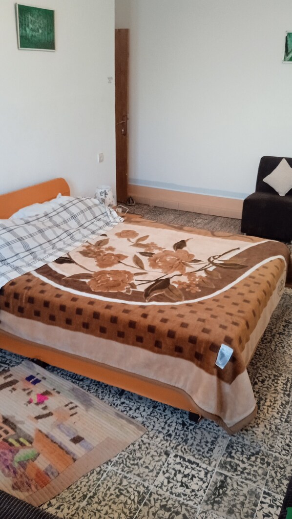 A bed with a floral bedspread, a patterned rug, and a chair in a room with plain walls and a wooden door. - welcomehome (Chefchaouen)