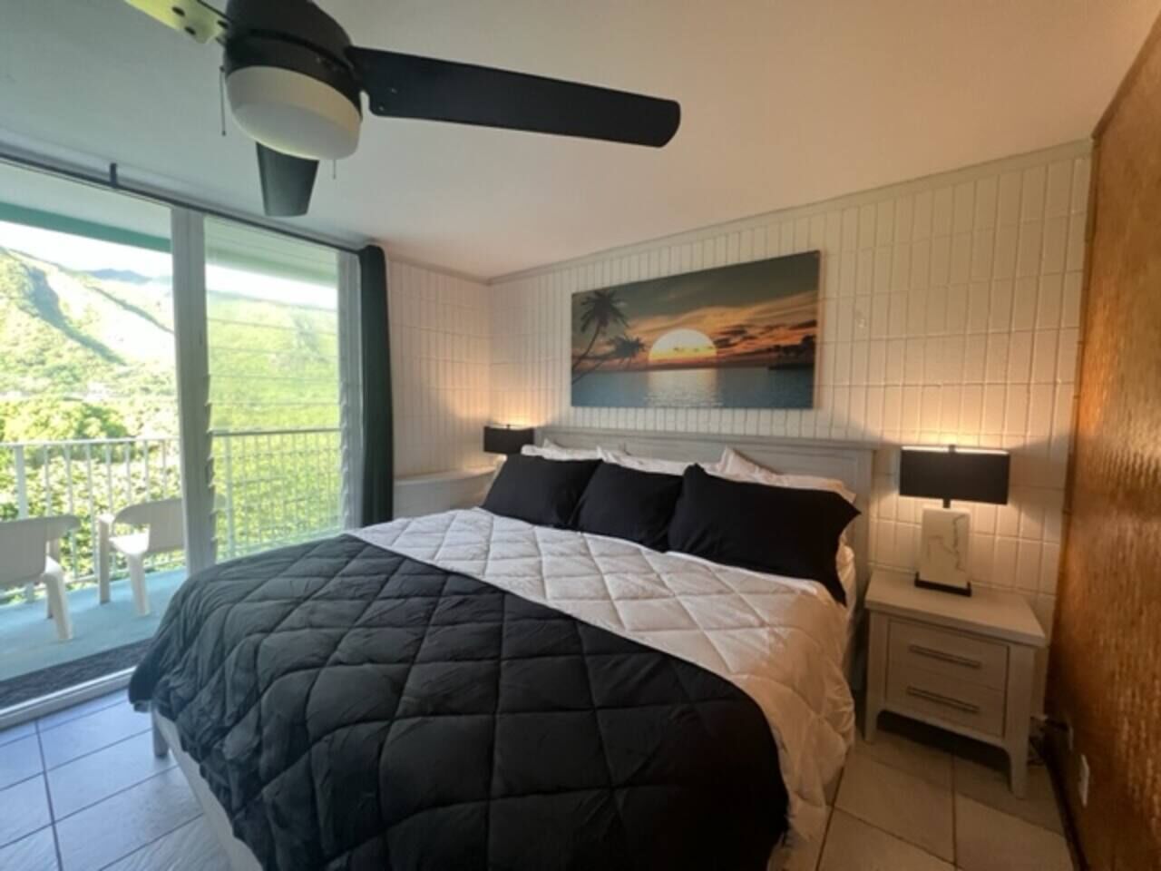 A bedroom with a large bed, a ceiling fan, a painting of a sunset, a balcony with chairs, and a nightstand with a lamp.