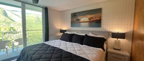A bedroom with a large bed, a ceiling fan, a painting of a sunset, a balcony with chairs, and a nightstand with a lamp.