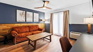 Habitació amb sòl de fusta, un sofà de cuir marró, taula de centre de fusta, ventilador de sostre, una làmpada, un radiador i una finestra amb cortines.