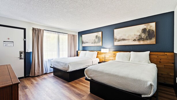 Una habitació d'hotel amb dos llits, un escriptori de fusta, una finestra gran amb cortines i dos quadres a la paret.