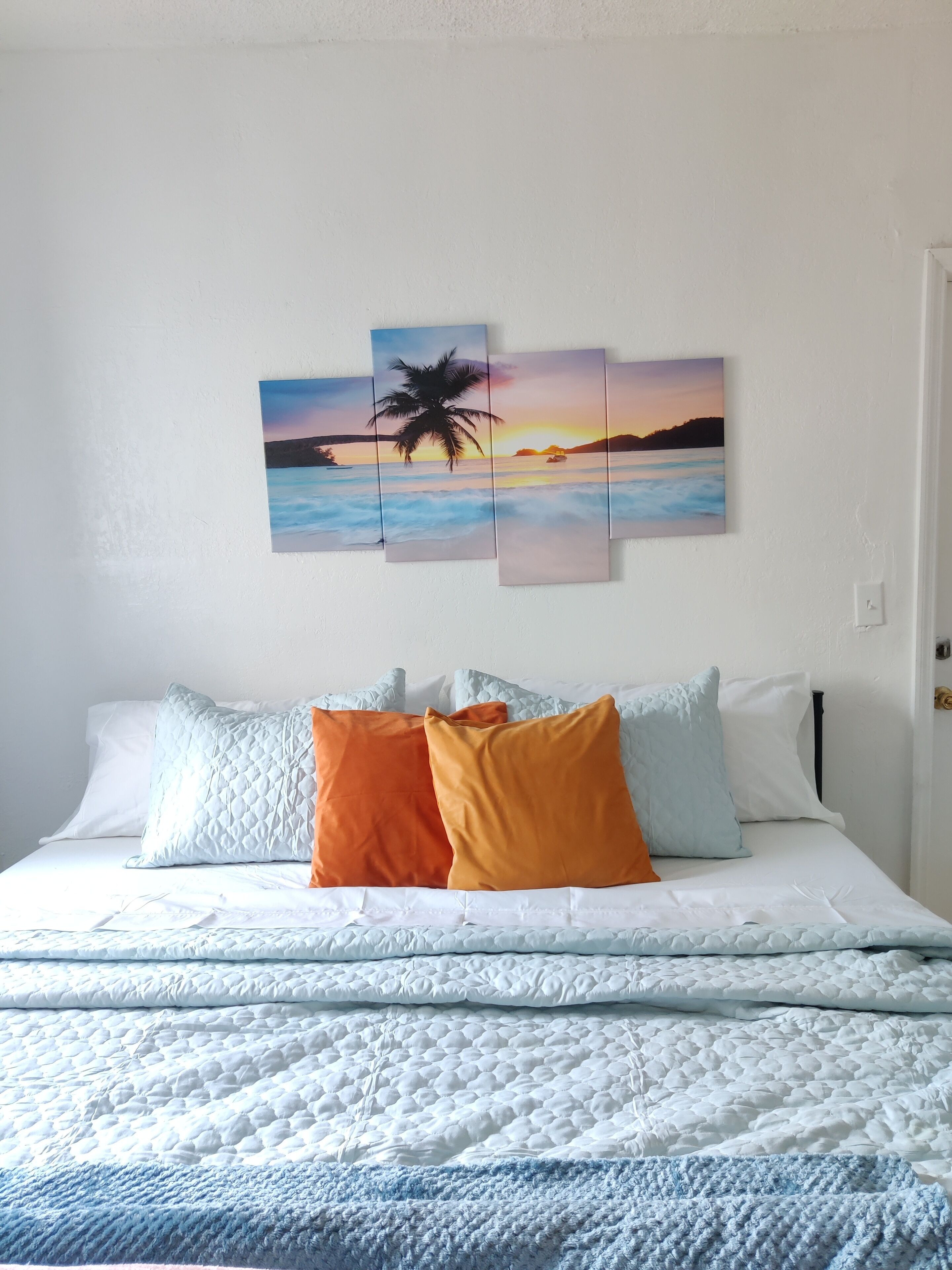 A bedroom with a bed, pillows, and a wall-mounted painting of a sunset over water.