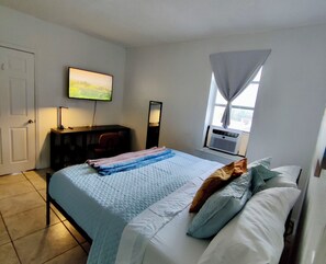Ein Schlafzimmer mit einem Bett, einem Fernseher an der Wand, einem Schreibtisch und einem Fenster mit Vorhängen.
