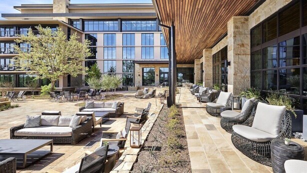 A modern outdoor lounge area with wicker furniture and stone flooring, overlooking a building with large windows. - King Room at a 5⭐️ Hotel (Frisco)