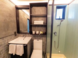 Un baño con lavamanos, inodoro y ducha. Hay repisas con toallas y artículos de aseo.