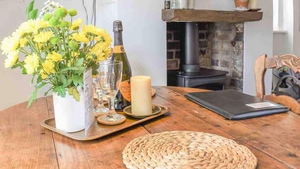 A wooden table with a woven placemat, a vase of yellow flowers, a candle, and a bottle of wine. - The Pepper Pot Sleeps 4 Sunlit Garden (Exmouth)