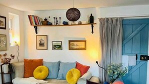 A cozy living room with a sofa, armchairs, and a coffee table. There is a bookshelf with books and decorative items, and a window with curtains. - The Pepper Pot Sleeps 4 Sunlit Garden (Exmouth)