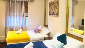Habitación de hotel con dos camas, una con cubrecama amarillo y la otra con cubrecama azul. Hay un espejo grande en la pared, un cuadro y una ventana con cortinas.