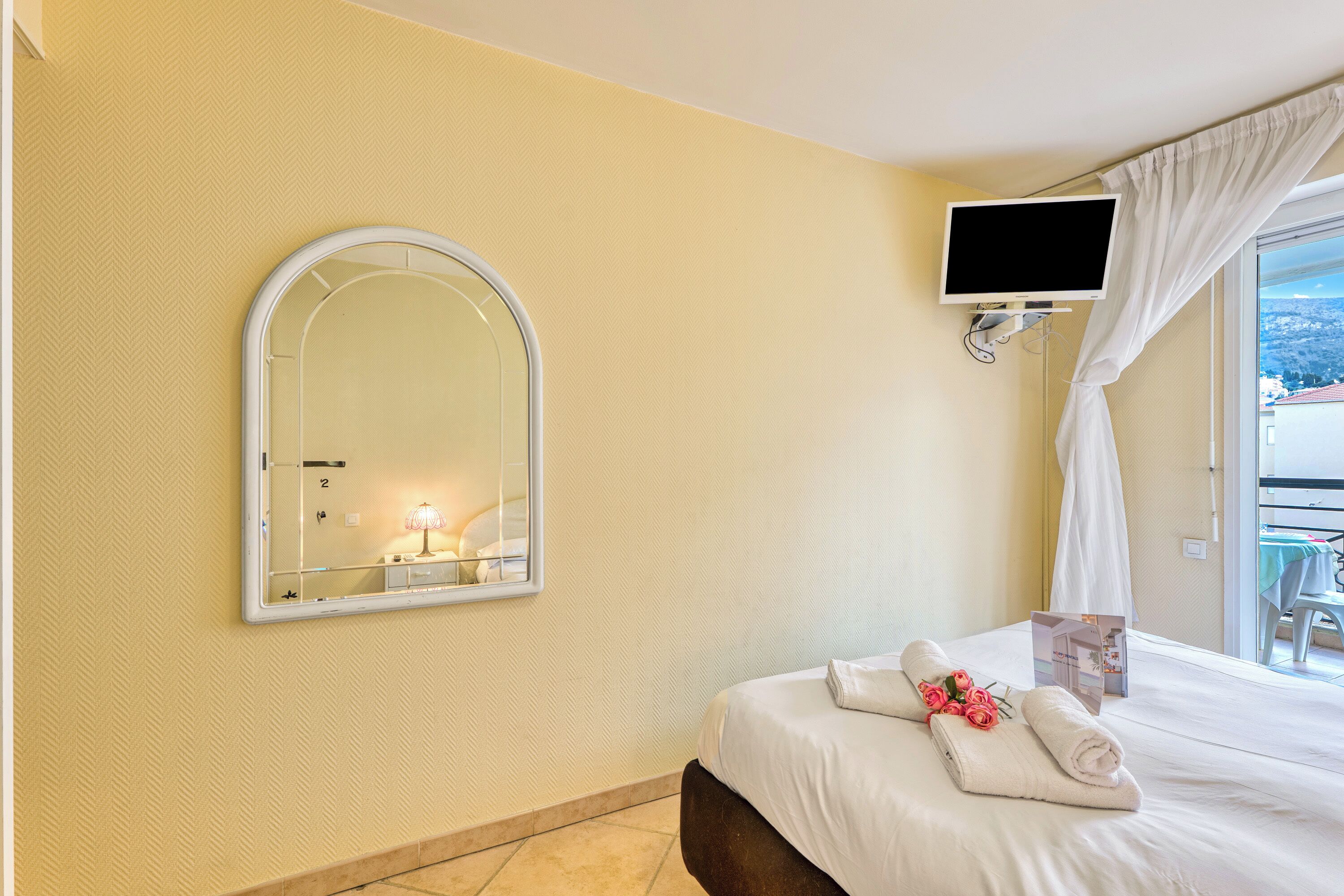 A hotel room with a bed, a television, a mirror, and a window with curtains.