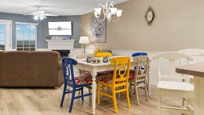 A dining area with a white table, colorful chairs, a chandelier, and a television on the wall. - Freshly Updated Lake-Access Condo with Outdoor Pool, Mini Golf, Pickleball & Free Attraction Tickets (Kimberling City)