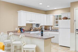 A kitchen with white cabinets, a central island with bar stools, a microwave, and a refrigerator. - Freshly Updated Lake-Access Condo with Outdoor Pool, Mini Golf, Pickleball & Free Attraction Tickets (Kimberling City)