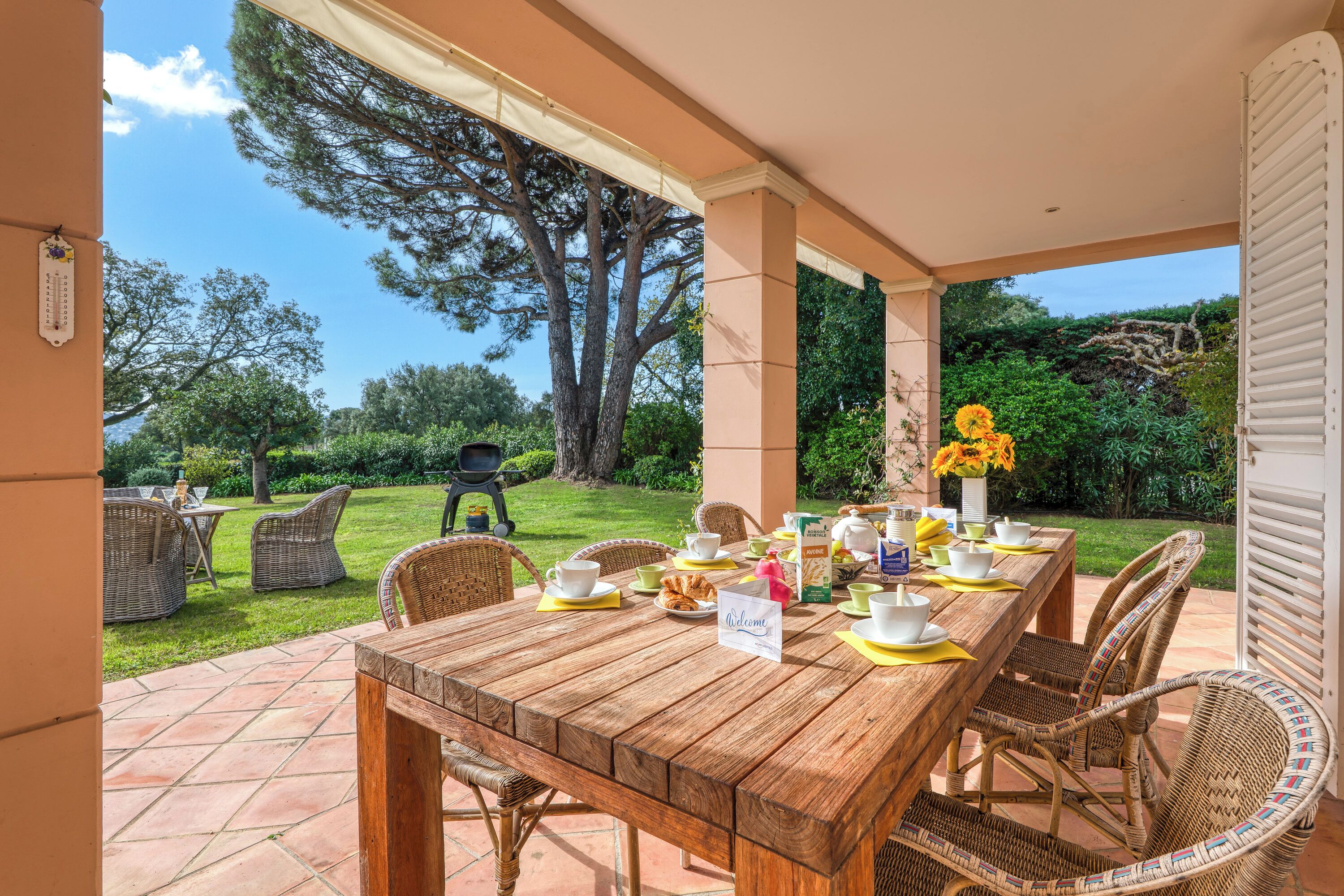 A wooden dining table set with chairs, a welcome sign, and a view of a garden with a barbecue grill.