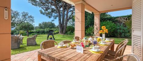 A wooden dining table set with chairs, a welcome sign, and a view of a garden with a barbecue grill.