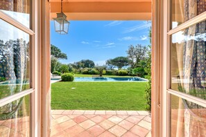 A view through a glass door leading to a well-maintained lawn, a swimming pool, and a clear blue sky. - Villa Tortue - Private pool & Sea view, Grimaud, France (Grimaud)