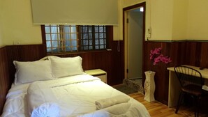 A neatly made bed with white linens, a wooden headboard, a small bedside table, a chair, and a window with blinds. - Dimocrata (Phnom Penh)