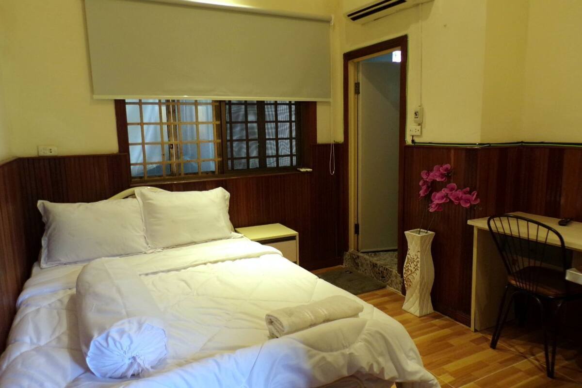 A neatly made bed with white linens, a wooden headboard, a small bedside table, a chair, and a window with blinds.