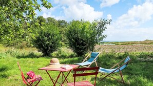 A red table with a straw hat and a book, two blue folding chairs, and a red folding chair in a grassy outdoor area with trees and a view of the sea.
