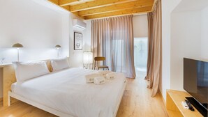 Een hotelkamer met een houten plafond, een bed met witte lakens, een nachtkastje met een lamp, een stoel, een televisie, en een zichtbare badkamer door een glazen deur.