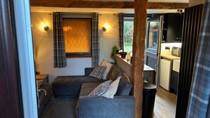 A cozy room with a sofa, a small table, a window with curtains, and a view of a kitchen area. - Quaint and Cosy Cottage - Manchester (Bury)