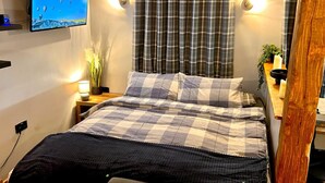 A neatly made bed with a checkered bedspread, a bedside lamp, and a wall-mounted TV displaying a weather forecast. - Quaint and Cosy Cottage - Manchester (Bury)