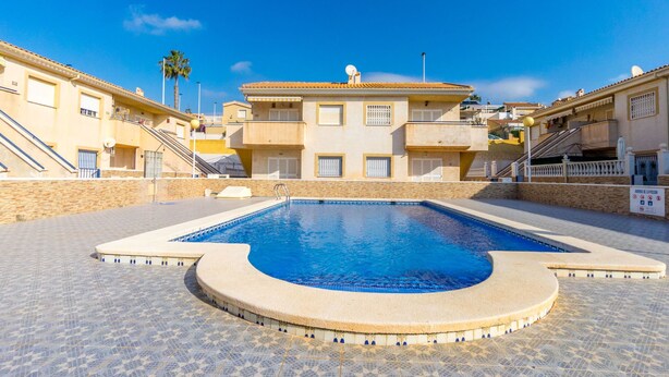 Una piscina con borde de baldosas, rodeada de edificios con balcones y un cielo azul despejado.