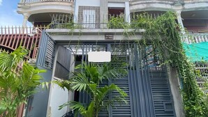 Un edificio de dos pisos con una entrada protegida por una reja, una planta en maceta al frente y un cielo despejado de fondo.