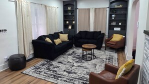 En moderne stue med en sort sofa, en brun lænestol og et lille træbord. To sorte hylder prydet med dekorative genstande og en loftventilator.