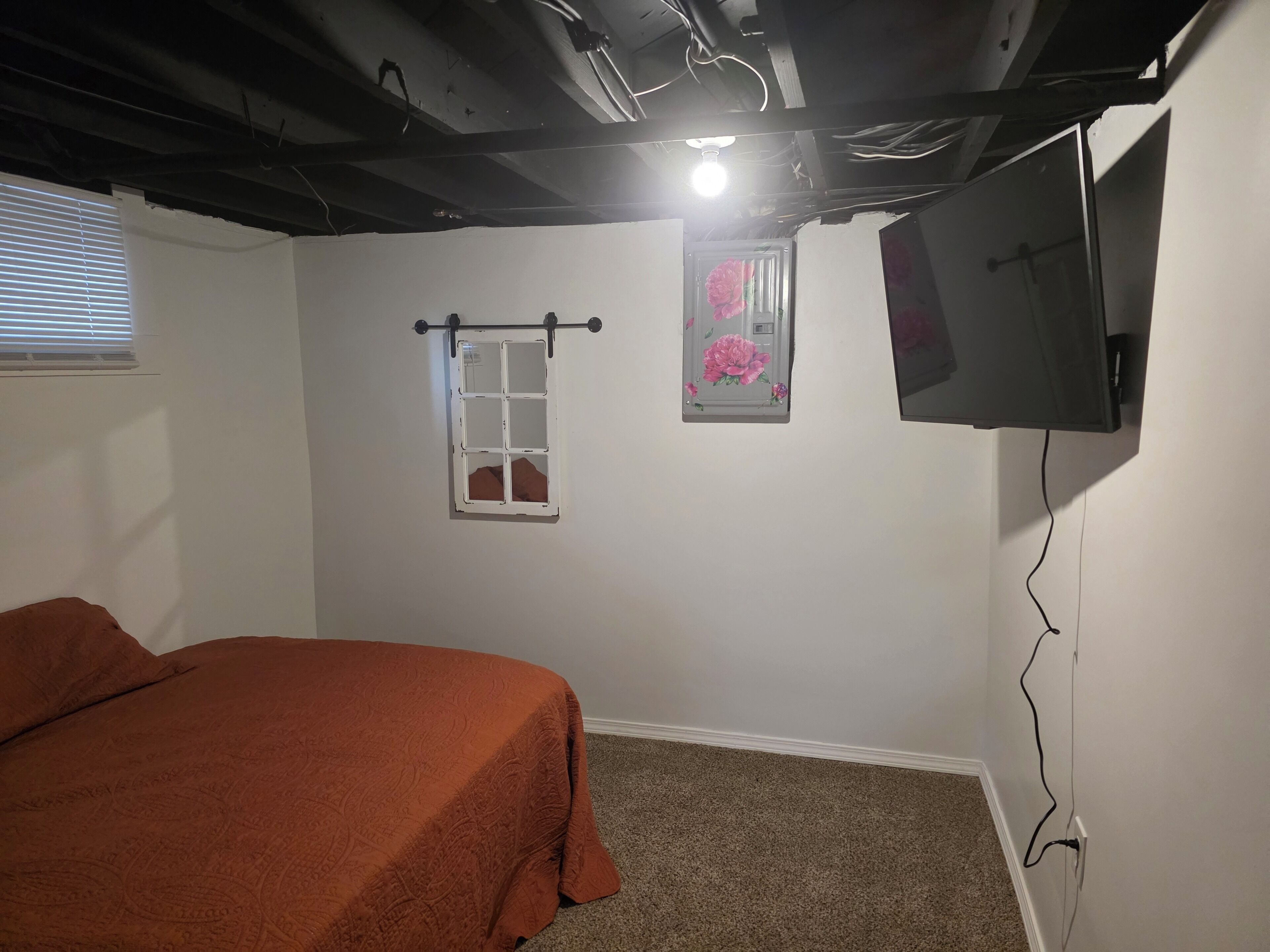 A bedroom with a bed, a television mounted on the wall, a window with blinds, and a small decorative item on the wall.