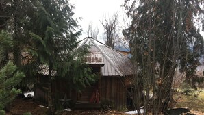 A rustic wooden cabin with a metal roof, surrounded by dense trees and a stone wall. - Cabin in Mountain Station - Easy Walk to Town and bordering the Rail to Trail (Nelson)