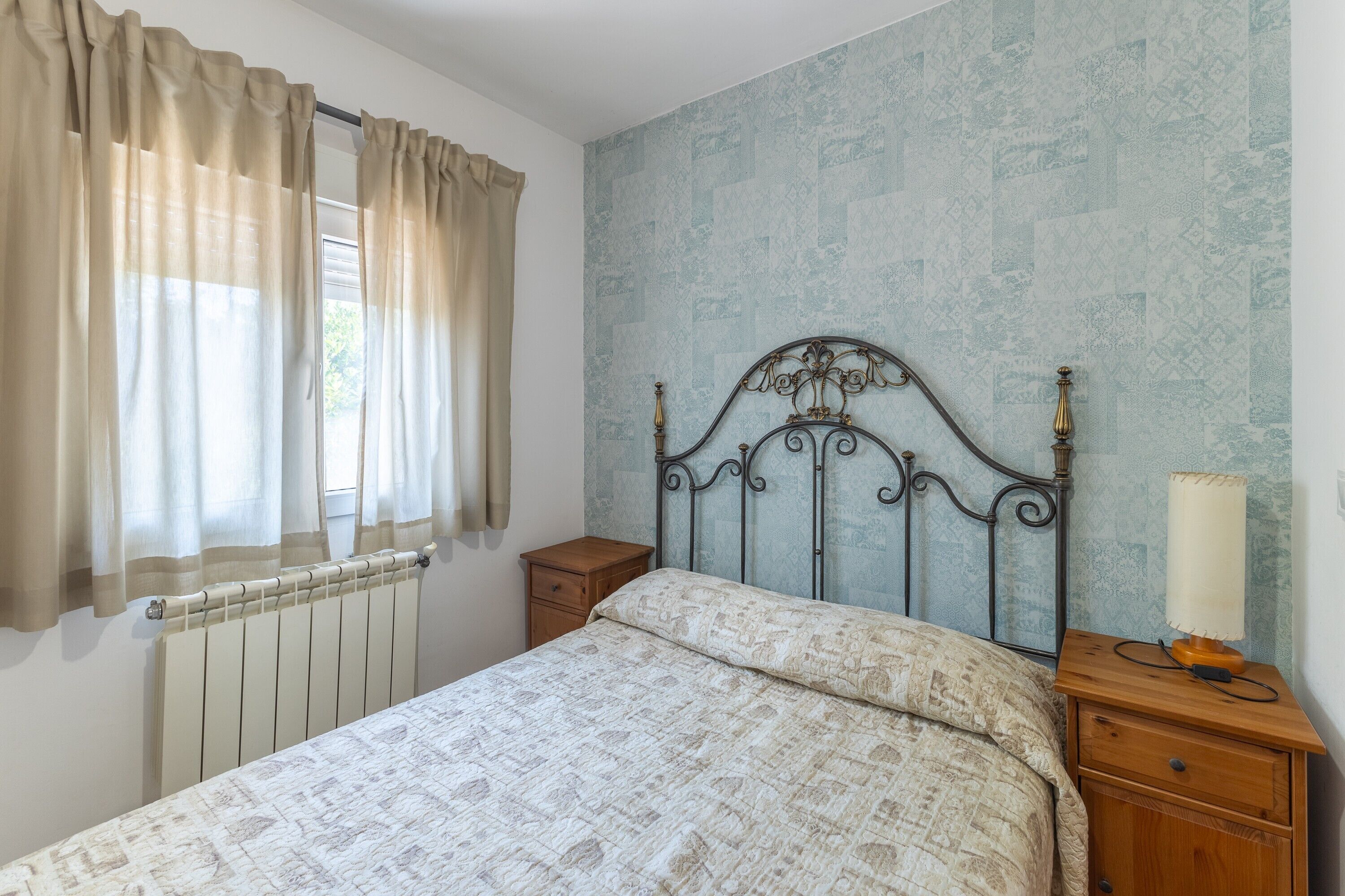 A bedroom with a bed, a nightstand, a lamp, a window with curtains, and a radiator.