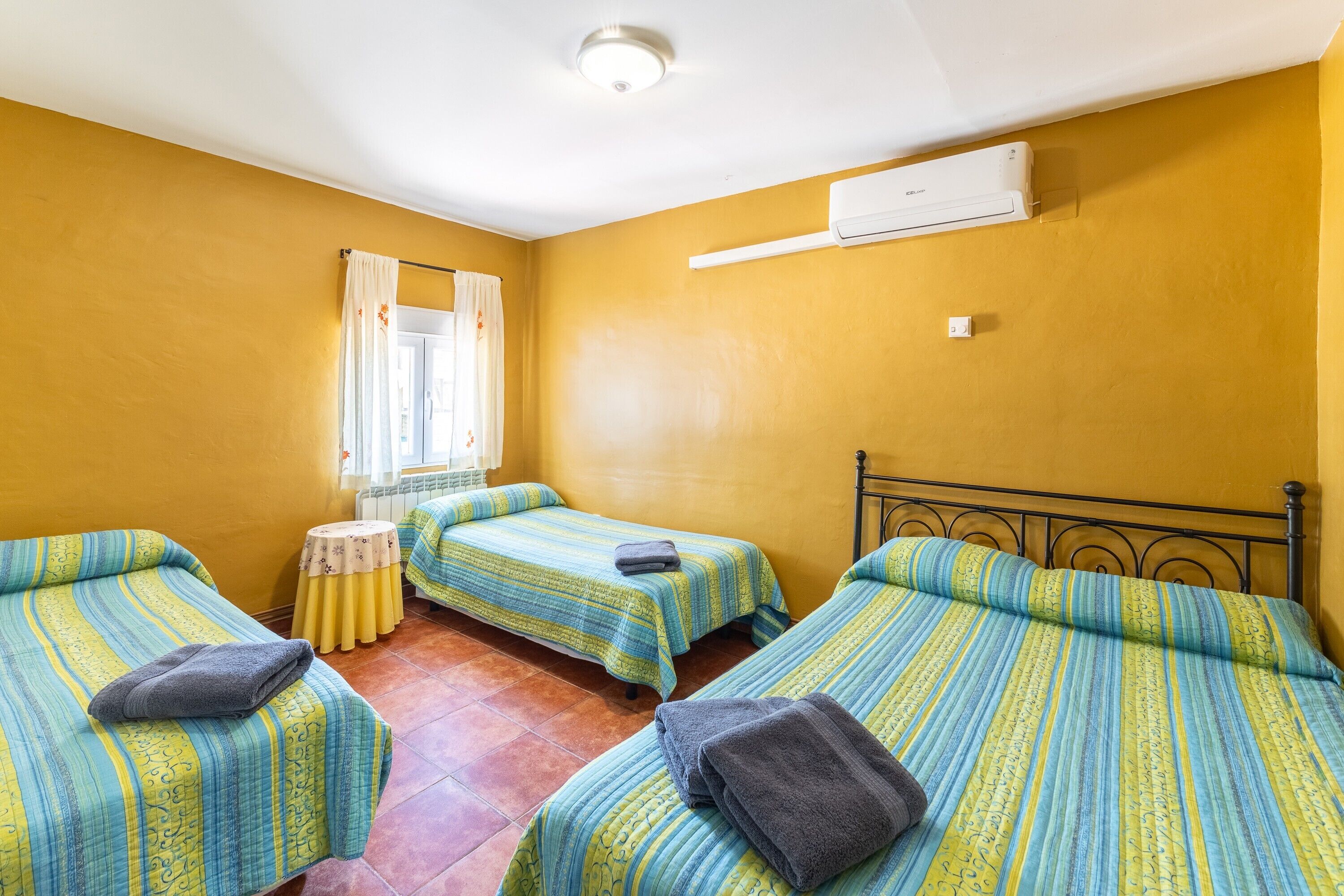 Una habitación con tres camas, una mesita redonda pequeña, un ventanal con cortinas y un equipo de aire acondicionado.