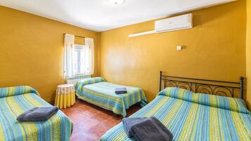 Una habitación con tres camas, una mesita redonda pequeña, un ventanal con cortinas y un equipo de aire acondicionado.