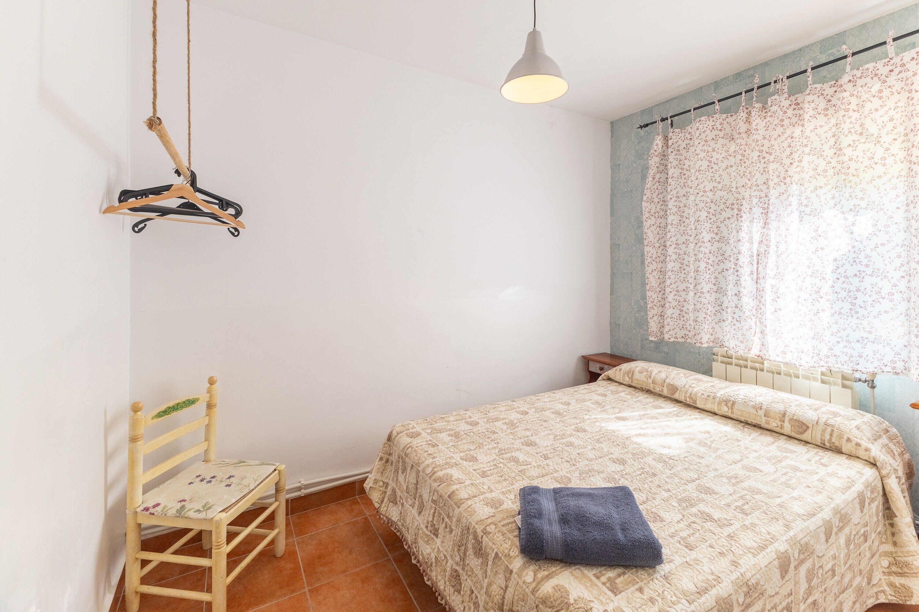 Een slaapkamer met een bed, een stoel, een hanglamp en een raam met gordijn.