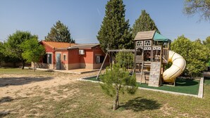 Un parque infantil con un tobogán, un árbol pequeño y un edificio de techo rojo.