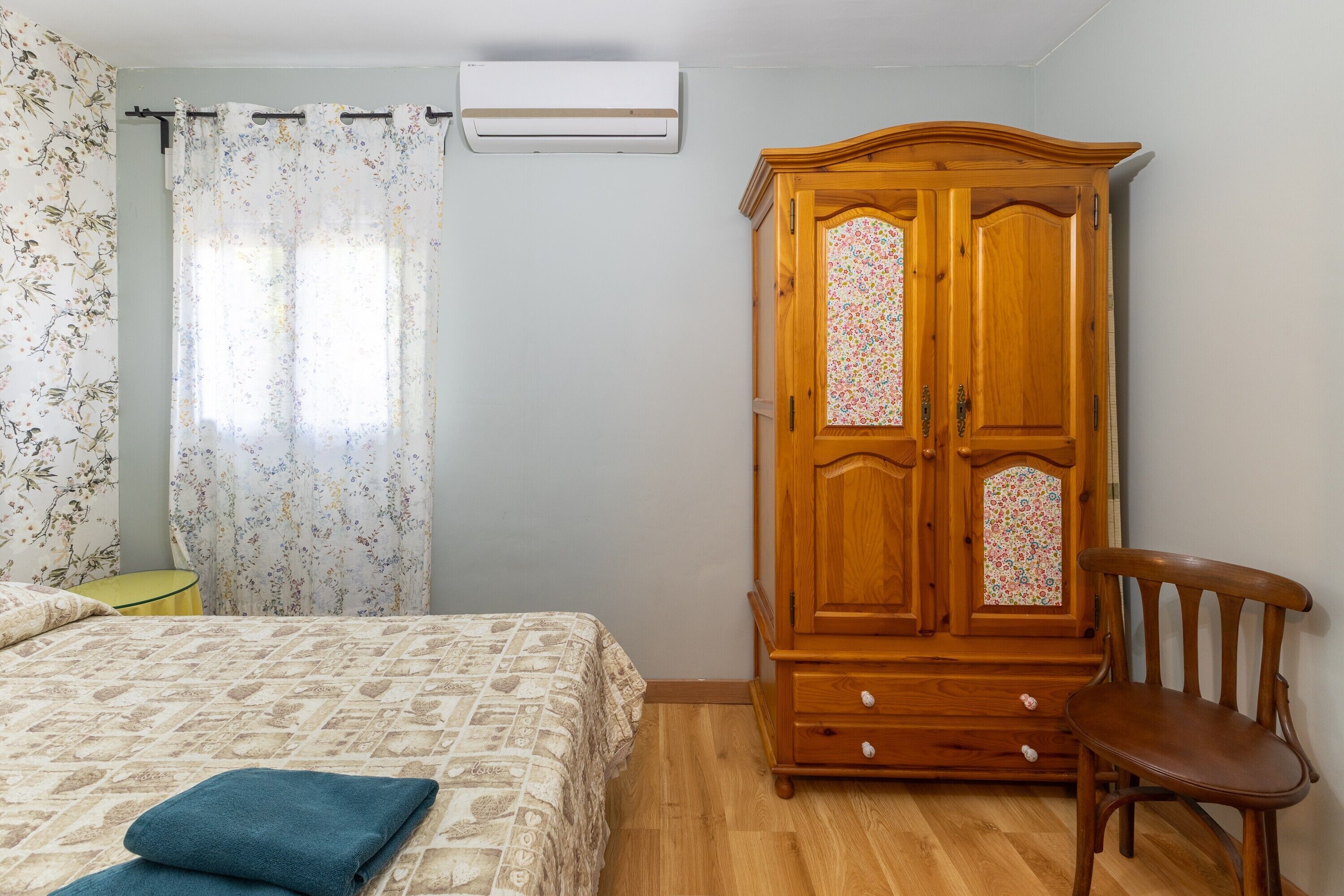 A bedroom with a bed, a wooden wardrobe, a chair, and a window with floral curtains.