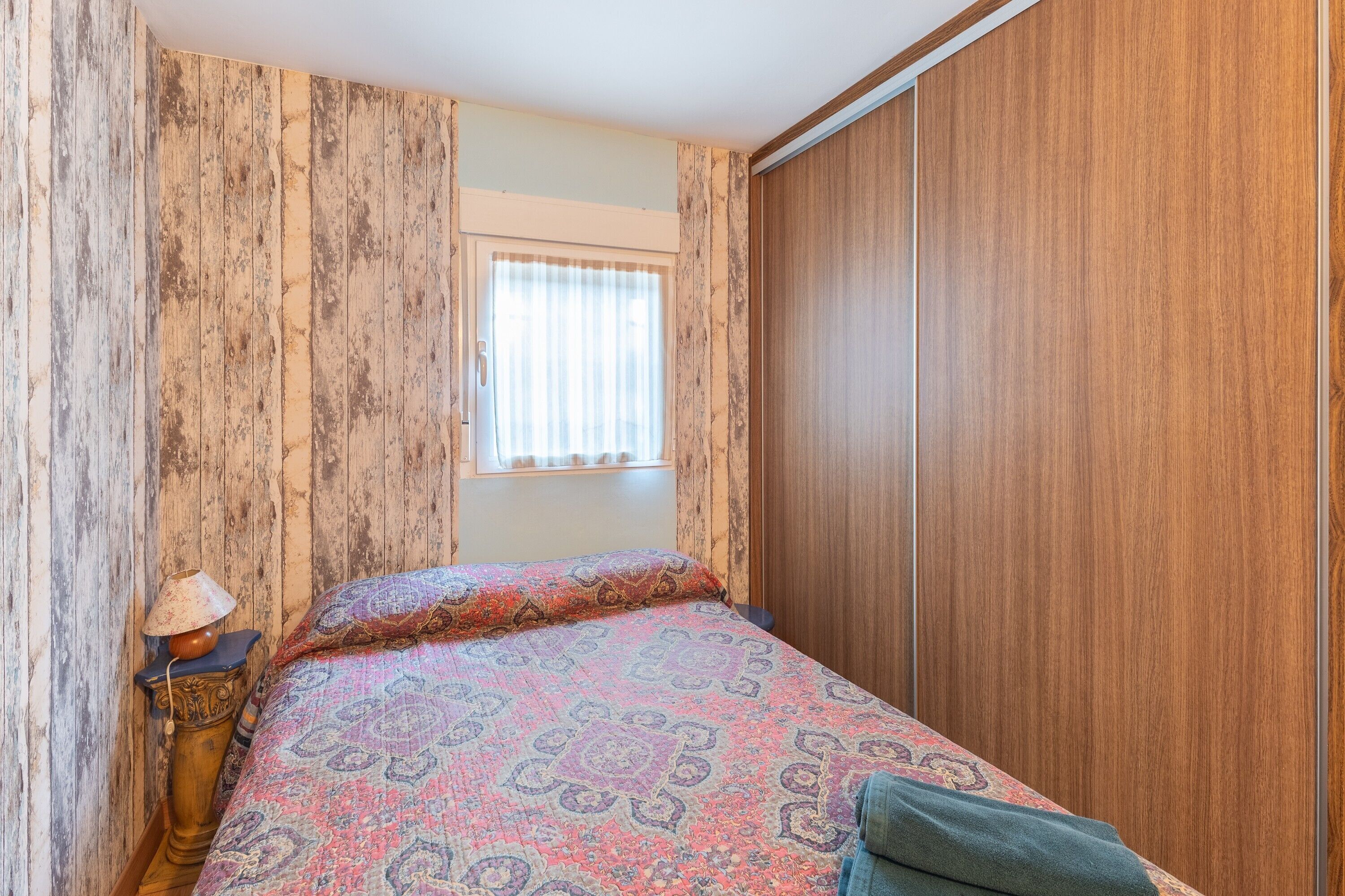 Une chambre à coucher avec une couvre-lit à motifs, une fenêtre avec des rideaux, une armoire en bois et une table de chevet avec une lampe.