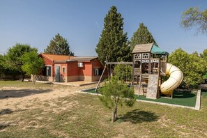 Une aire de jeux avec un toboggan et des structures d’escalade, un petit bâtiment avec un toit rouge et une zone herbeuse avec des arbres.