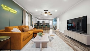 A modern living room with a sofa, coffee table, TV, and kitchen in the background. - Adventure Basecamp, Walks to Kaymoor and Long point Trails, Hot Tub & Game Room (Fayetteville)