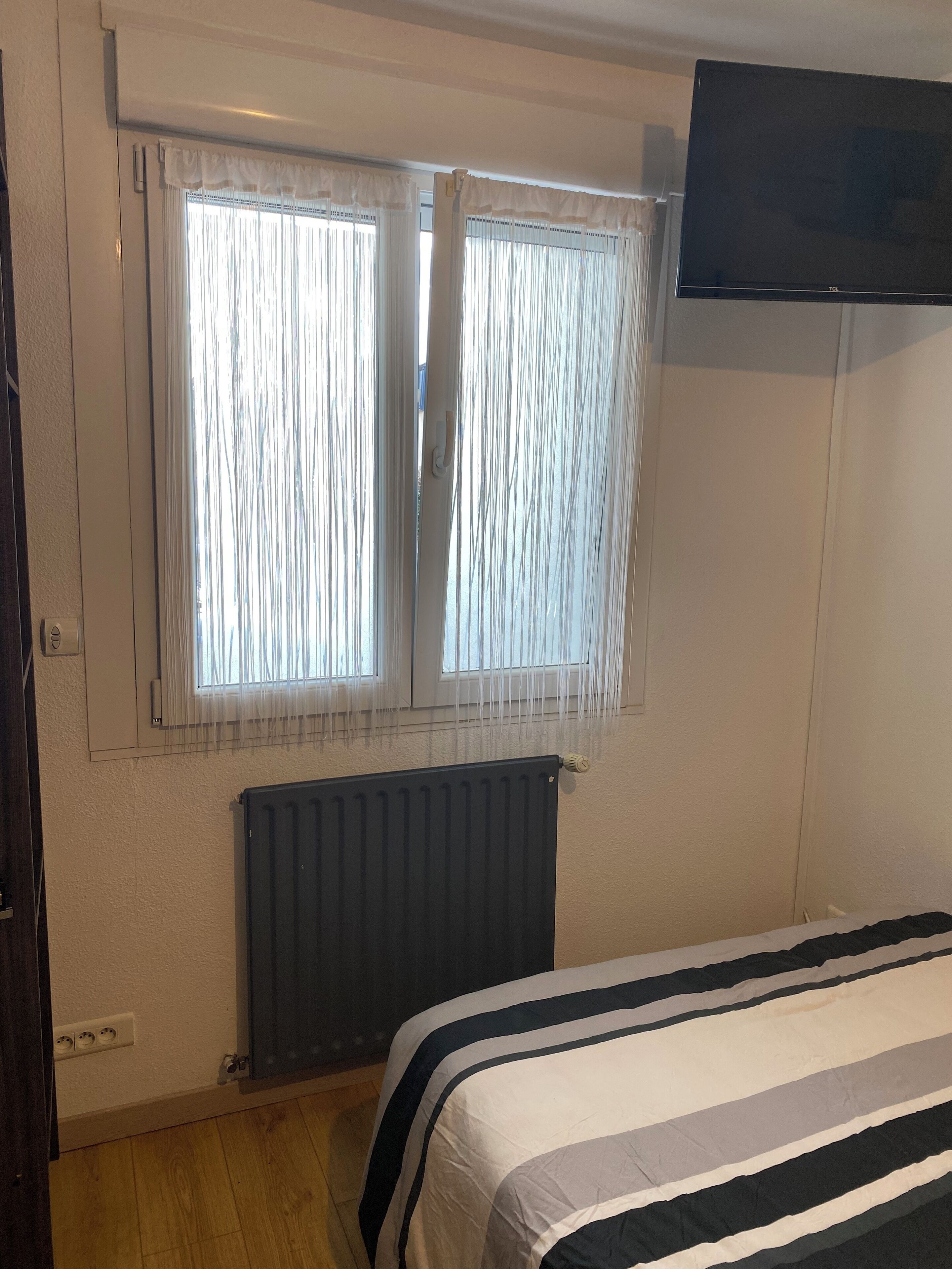 Un dormitorio con cama, dos ventanas con cortinas, un televisor montado en la pared y un radiador.