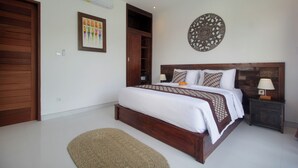A bedroom with a wooden bed, a nightstand with a lamp, a wall-mounted decorative plate, and a framed picture on the wall. - Bougainvillea Villas Cemagi by Parigata (Cemagi)