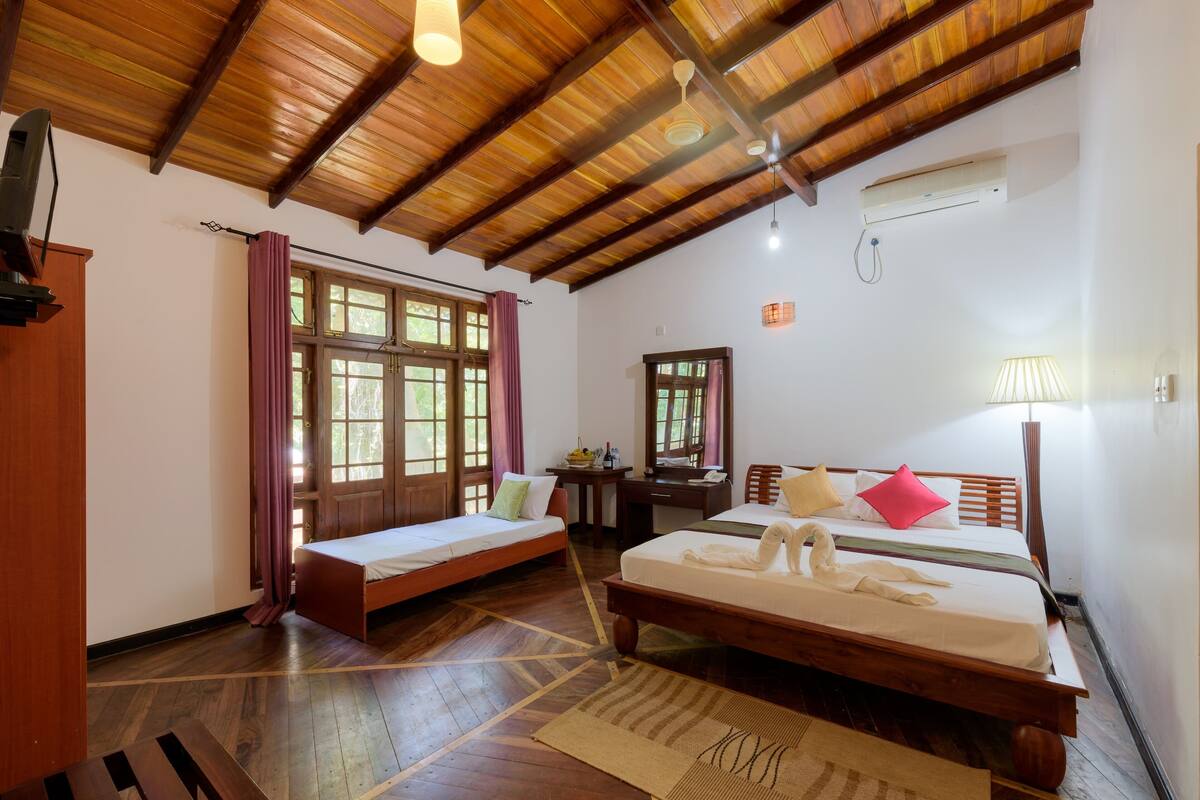 A room with two beds, wooden flooring, a ceiling fan, an air conditioning unit, and a window with curtains.